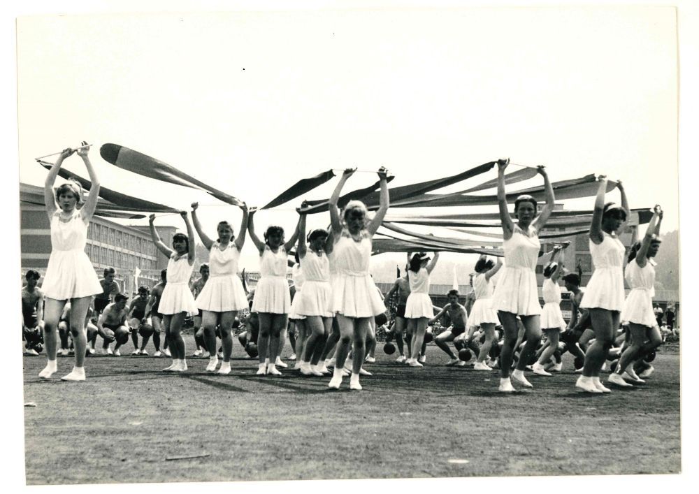 Fotografie pravděpodobně z roku 1959 zachycuje atmosféru okresní spartakiády v Rožnově pod Radhoštěm, jež byla přípravou na celostátní spartakiádu v následujícím roce. Snímek, který je jinak datován (snad chybně) do roku 1960 a jehož autora neznáme, odráží nejen sportovní a společenský rozměr spartakiád, ale i důraz na kolektiv, jednotu a estetickou působivost, které byly pro spartakiádní hnutí charakteristické. Státní okresní archiv Vsetín, fond Sbírka fotografií, č. 2860.